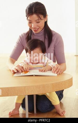 Vierzehn Monate altes Baby Mädchen am Tisch, auf dem Schoß der Mutter Lesung Buch sitzen Stockfoto