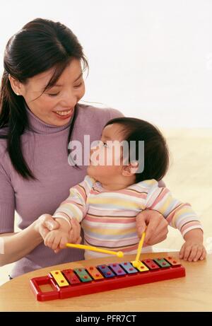 Vierzehn Monate altes Baby Mädchen sitzt auf dem Schoß der Mutter spielen mit Spielzeug Xylophon Stockfoto