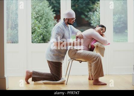 Schwangere Frau auf Stuhl sitzt in ihrem Rücken von Mann massiert. Stockfoto