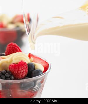 Pouring cream from a jug onto summer fruit including raspberries, blueberries, blackberries and strawberries, close-up Stockfoto