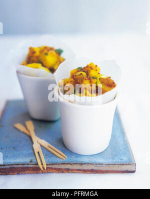 Zwei Tassen mit Kartoffeln mit Kurkuma und Senf Samen (Aloo chaat) und zwei hölzerne chip Gabeln, auf blauen Keramikfliesen Stockfoto