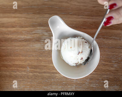 Schaufel mit hausgemachten Rum Rosinen Eis mit Edelstahl Löffel auf weibliche Hand in kleinen weißen Schale auf Holz- Hintergrund Stockfoto