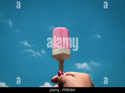 Woman's Hand mit roten Nägeln Holding Pink Popsicle Eis gefroren Pop auf Sommer und blauer Himmel Stockfoto
