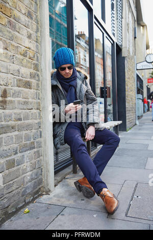 Junger Mann auf einer Bank sitzen außerhalb, auf sein Handy zu schielen. Stockfoto