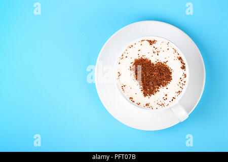 Schließen Sie eine volle weiße Tasse cremiger Latte Cappuccino Kaffee mit Schokolade in Herzform Kunst in Pastell-blaue Papier Hintergrund, erhöhte Ansicht von oben, di Stockfoto