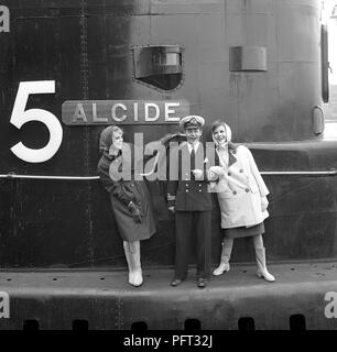 60er Jahre Mode. Zwei Models und der Kapitän der Britischen u-boot Alcide. Das U-Boot liegt im Hafen von Stockholm verankert. Die Modelle tragen die Herbste mode Mäntel und Schals. Schweden Mai 1962 Stockfoto