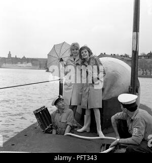 60er Jahre Mode. Zwei fashion Modelle und die Besatzung der Britischen u-boot Alcide. Das U-Boot liegt im Hafen von Stockholm verankert. Die Modelle tragen die herbste Mode Der regenjacken. Schweden Mai 1962 Stockfoto