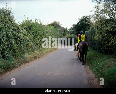 Zwei junge Frauen mit Leuchtstoff tops reiten Ponys auf Feldweg, Rückansicht mit Blick auf den Gegenverkehr Stockfoto