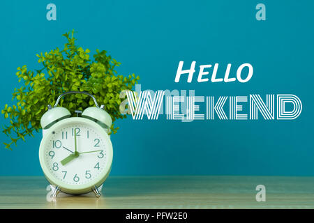 Hallo Wochenende Text auf grünem Hintergrund und kleinen Baum in weiße Vase mit Vintage Wecker auf Holztisch. Stockfoto
