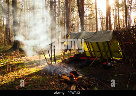 Bushcraft survival Tierheim Campingplatz im Wald Wildnis mit Rauch- und Sonnenstrahlen. Tarp/Zeltlager im Wald mit Lagerfeuer außerhalb. Stockfoto