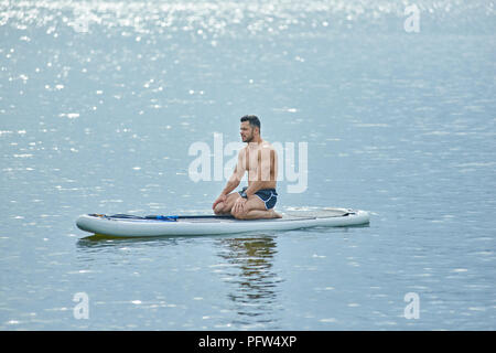 Sportliche Mann sitzt auf sup Board während trainining auf See, freuen. In gesund, muskulösen Körper. Lange oar Festlegung in der Nähe, suop Board schwimmen auf blaues Wasser Oberfläche. Warme, sonnige Wetter. Stockfoto