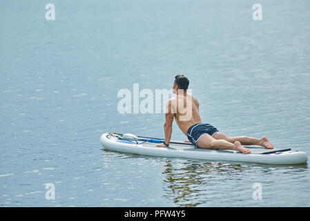 Passen Mann tun Dehnübungen Verlegung auf sup Board, Schwimmen auf dem See Training während der sonnigen Sommertag. Männliches Modell in Passform, muskulöse Körper, die Aufrechterhaltung gesunder Lebensstil. Stockfoto