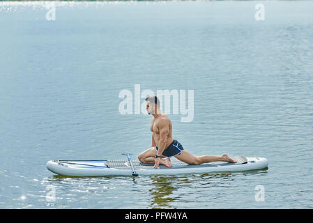 Seitenansicht von fit Mann zu tun Stretching Übungen sitzen auf sup Board, Schwimmen in Stadt See während der sonnigen Sommertag. Männliches Modell in sportlich fit Körper mit starken Muskeln. Lange oar Festlegung in der Nähe. Stockfoto