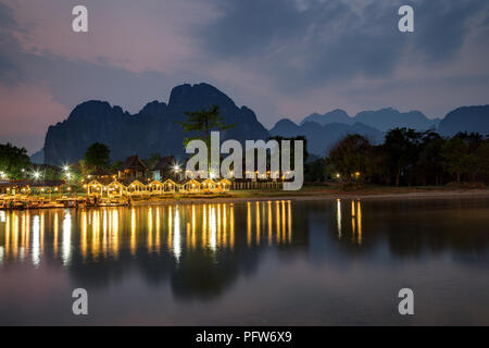 Nur wenige Menschen an einer beleuchteten Restaurant Waterfront durch den Nam Song Fluss und die Silhouette von Karst Kalkstein Berge in Vang Vieng, Laos, in der Dämmerung. Stockfoto