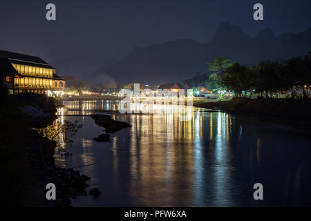 Silhouette von Karst Kalkstein Berge und Reflexionen von beleuchteten Restaurants am Wasser vom Fluss Nam Song in Vang Vieng, Laos, bei Nacht. Stockfoto