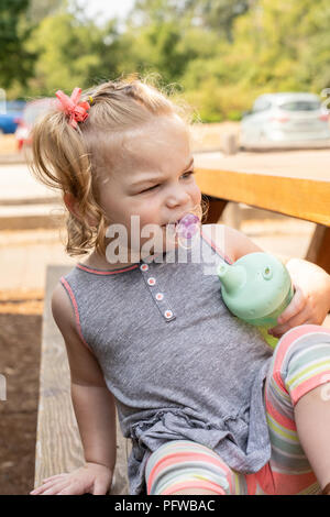 20 Monate altes Mädchen an einem Picknicktisch sitzen, während Ihr sippy cup Holding, suchen sehr in Gedanken verloren Stockfoto