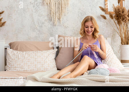 Eine junge Frau in einem lila Home Kleid strickt Ein hellrosa Schal oder Plaid aus natürlichen Threads auf dem Bett, gegen eine weiße Wand Stockfoto