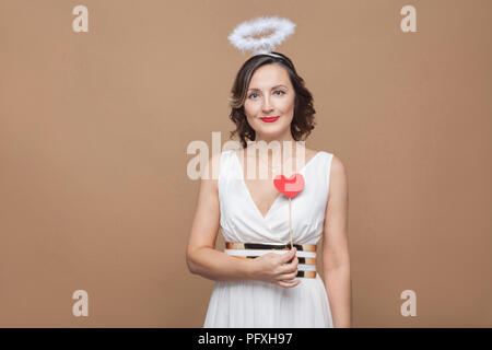 Mitte agaed Engel elegante brunette Frau in weißem Kleid mit Nimbus auf dem Kopf halten roten Herzen Sticker und Kamera mit Lächeln. Studio shot, Stockfoto