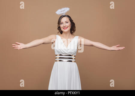 Engel im mittleren Alter elegante brunette Frau in weißem Kleid will zu umarmen und Kamera mit toothy Lächeln und große erhobenen Arme. Studio shot, Hallenbad Stockfoto