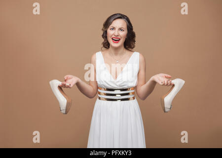 Wütend business Frau in weißem Kleid auf Kamera suchen, die schreien und Holding unbequem High Heels. Emotion und Gefühl Konzept. Studio sho Stockfoto