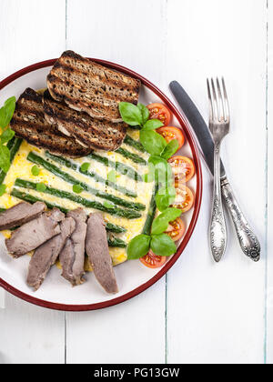 Frittata mit Spargel und Basilikum. Hausgemachten Schinken, Tomaten, Toast. Holz- hellen Hintergrund. Vertikaler Dampfstoß Stockfoto