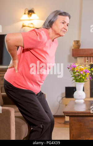 Reife Frau leiden unter Rückenschmerzen zu Hause. Unterer Rücken Massage mit Hand, das Gefühl erschöpft, stehend im Wohnzimmer. Stockfoto