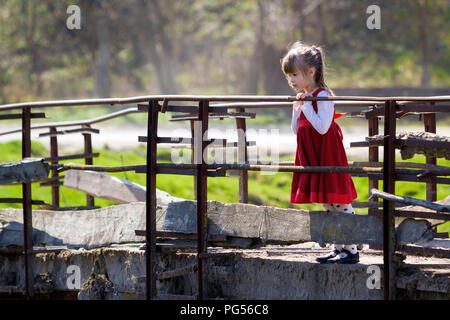 Die hübsche kleine Blonde langhaarige Mädchen in schönes rotes Kleid steht allein auf alten Zement Brücke gelehnt auf hölzernen Geländer versessen auf warmen sonnigen Stockfoto