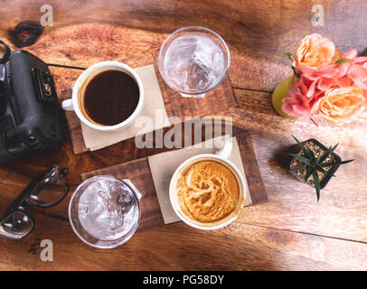 Ansicht von oben schwarz Kaffee und Milch Kaffee in weisser Keramik Schale auf Holz- Hintergrund mit Blume und Retro Gläser und Eiswasser. Stockfoto