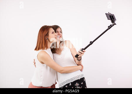 Porträt von zwei schöne junge Frau eine selfie mit einem selfie Stick und andere lustig und dumme Gesichter, während es zu tun. Studio Fotos über eine weiße Wand Stockfoto