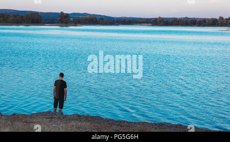 Junge Mann stand und zurück mit legere Kleidung, zurück an den Ufern des Blauen Sees bei Sonnenuntergang, mit Eukalyptus Wald im Hintergrund, Spanien Stockfoto