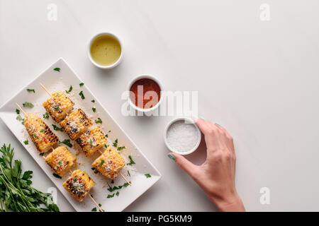 7/8 Bild der Frau Salz aus Marmor tisch mit gegrilltem Mais auf Platte in der Nähe von Petersilie Stockfoto