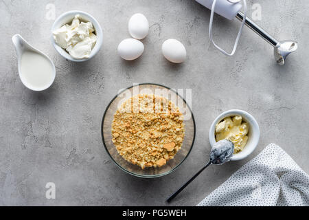 Flach mit zerquetschten Cookies, rohem Huhn Eier und Butter für die Torte auf graue Oberfläche Stockfoto