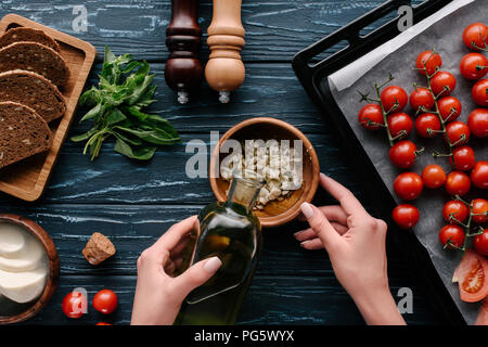 7/8-Ansicht von weiblichen Händen Hinzufügen von Öl auf dunklen Holztisch mit Tomaten und Kräutern, Knoblauch Stockfoto