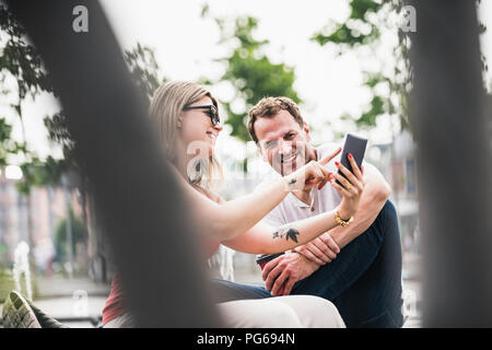 Glückliches Paar auf einer Bank sitzen teilen Handy Stockfoto