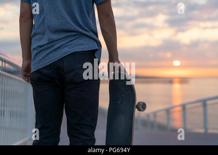 Junger Mann mit Skateboard am Strand bei Sonnenaufgang, Rückansicht Stockfoto