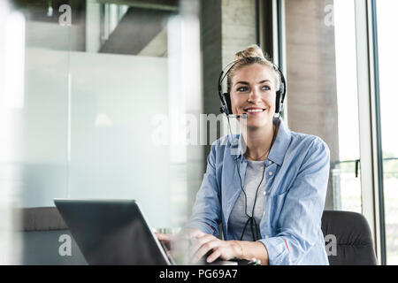 Junge geschäftsfrau am Schreibtisch sitzend, einen Anruf zu tätigen, die mit dem Headset und Laptop Stockfoto