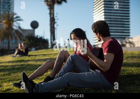 Spanien, Barcelona, junger Mann mit Gitarre für Freundin sitzen auf einer Wiese Stockfoto
