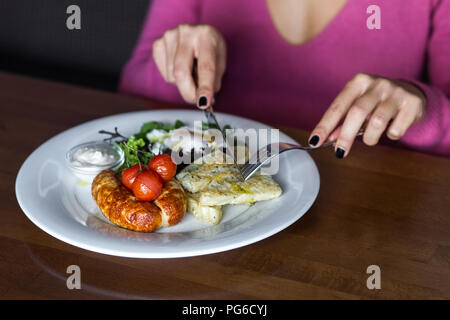 Frühstück mit gebratenen Würstchen, baked Cherry Tomaten, pochierte Eier und Grüns. Frau mit Messer und Gabel Stockfoto