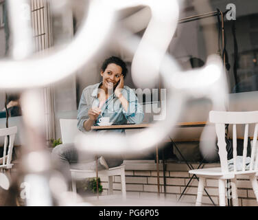 Junge Frau trinkt Kaffee vor Cafe, am Telefon zu sprechen Stockfoto