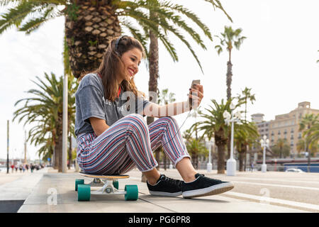 Glückliches junges Mädchen sitzt auf Skateboard Musikhören mit Kopfhörern Stockfoto