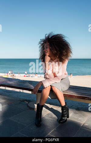 Schöne junge Frau mit Afro Frisur sitzen auf einer Bank am Strand Stockfoto
