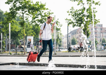 Mann mit rollenden Koffer und Handy zu Fuß am Brunnen in der Stadt Stockfoto