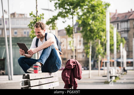 Mann mit Takeaway Kaffee sitzt auf der Bank mit Tablet Stockfoto