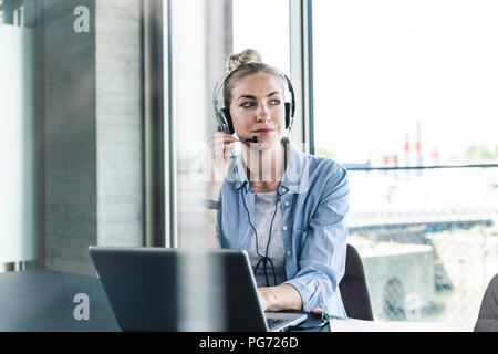 Junge geschäftsfrau am Schreibtisch sitzend, einen Anruf zu tätigen, die mit dem Headset und Laptop Stockfoto