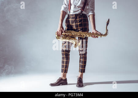 7/8 Schuß von stilvollen junge Musiker stehen und Holding Saxophon in Rauch auf Grau Stockfoto