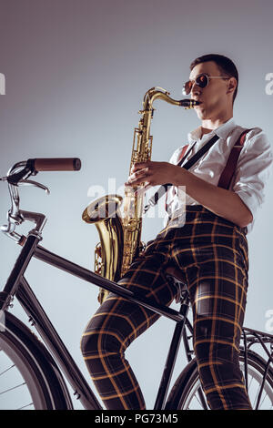 Low Angle View der jungen Schauspieler sitzen auf dem Fahrrad und spielt Saxophon auf Grau Stockfoto