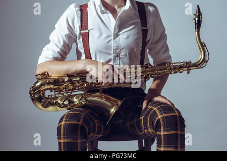 7/8 shot der jungen Musiker sitzt auf einem Stuhl und halten Saxophon isoliert auf Grau Stockfoto