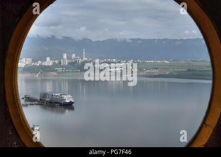 Blick über Yangtze Fluss durch das Runde Fenster. Ein Boot auf dem Fluss, die Berge und die kleine Stadt im Hintergrund. Stockfoto