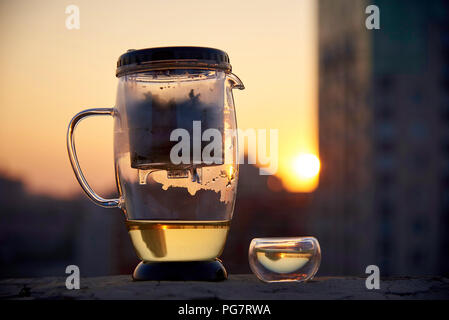 Eine Teekanne aus Glas und einer Tasse Tee auf einem unscharfen Hintergrund des Abends Stadt im Licht des Sonnenuntergangs. Stockfoto