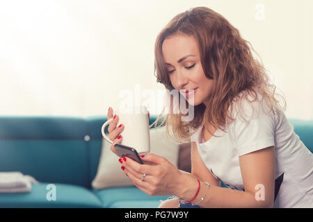 Junge Frau sitzt auf der Couch und Sie Ihr Smartphone während mit Tasse Kaffee bei Tageslicht Stockfoto